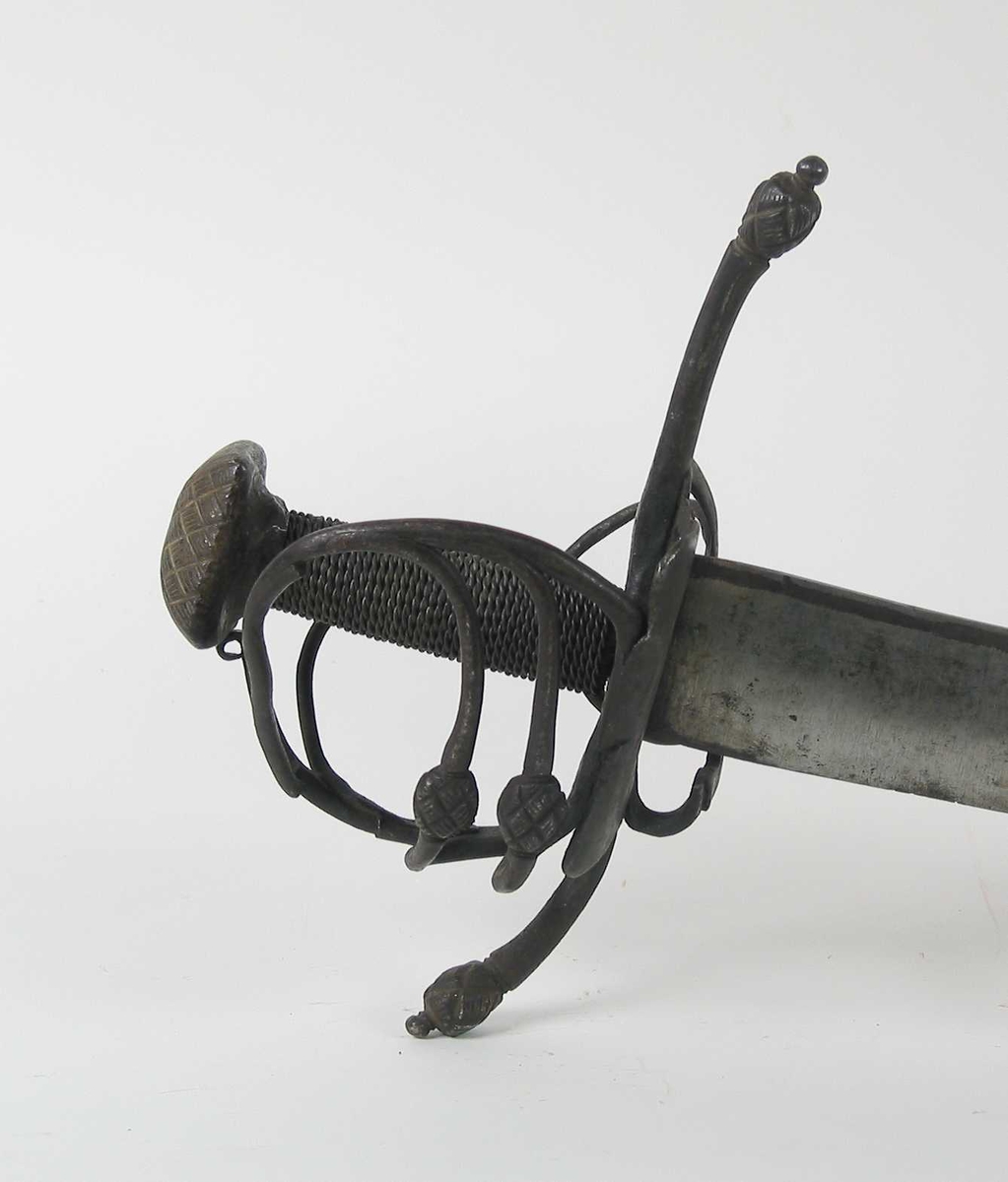 Pyramideformet knapp med strekdekor. Asymmetrisk kurvfeste med to sidebøyler( øverste avbrukket )med riflede fortykkelser på midten. mellom håndbøylen og kurvens ytterkant. Innsiden har en  sidebøyle. Kurvens basis er på utsiden en kvadratisk parérplate med to hjerteformede gjennombrytninger. På innsiden er det en klingebrekker og tommelbøyle. Opp og nedbøyde parérstenger med riflede fortykkelser på endene.
Tregrep med nyere jerntrådvikling.
Enegget krum klinge med 3 hull ipå klingspissen og en smal hulslipning på ryggsiden.Tre smedmerker på venstre side. 