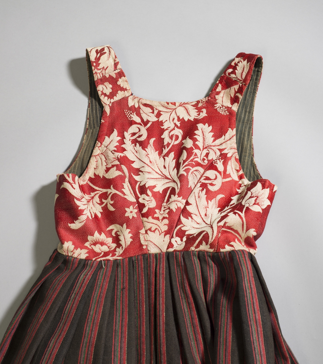 Håndsydd livkjole med stripete ullskjørt og rødt ullliv med store plantemotiv. Livet har rett hals bak, skjøtte seler og mangler ryggsøm. Lukningen består av fire par hekter og tre par hemper til snøring. Åpen klaff.
Fõr av stripete håndvevd ullstoff.
Nederst på skjørtet et børstebånd og skoning av et grindvevd bånd.
Skjørtet har en hengefald.