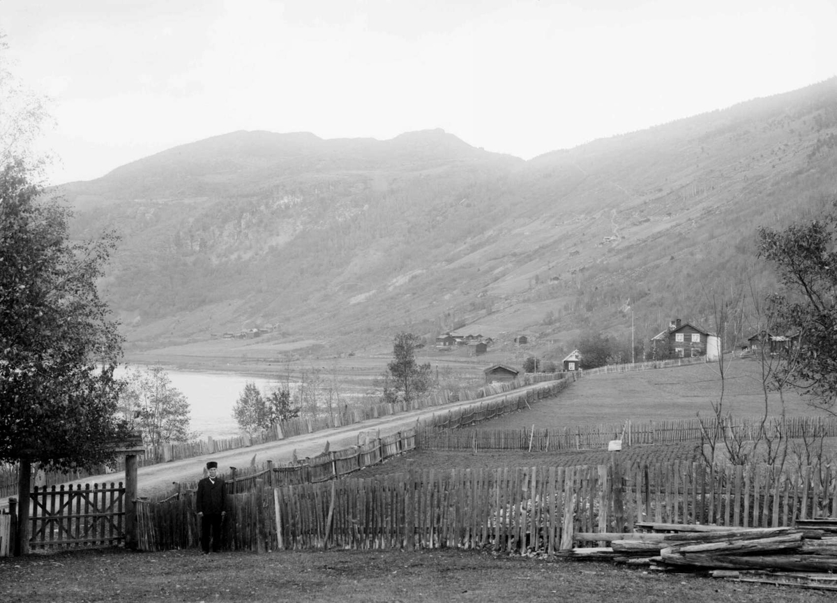 07.10.1909. Parti fra Bredebygden. Bebyggelse, vei, gjerder, mann ved port, åsside med bebyggelse og skog, elv.