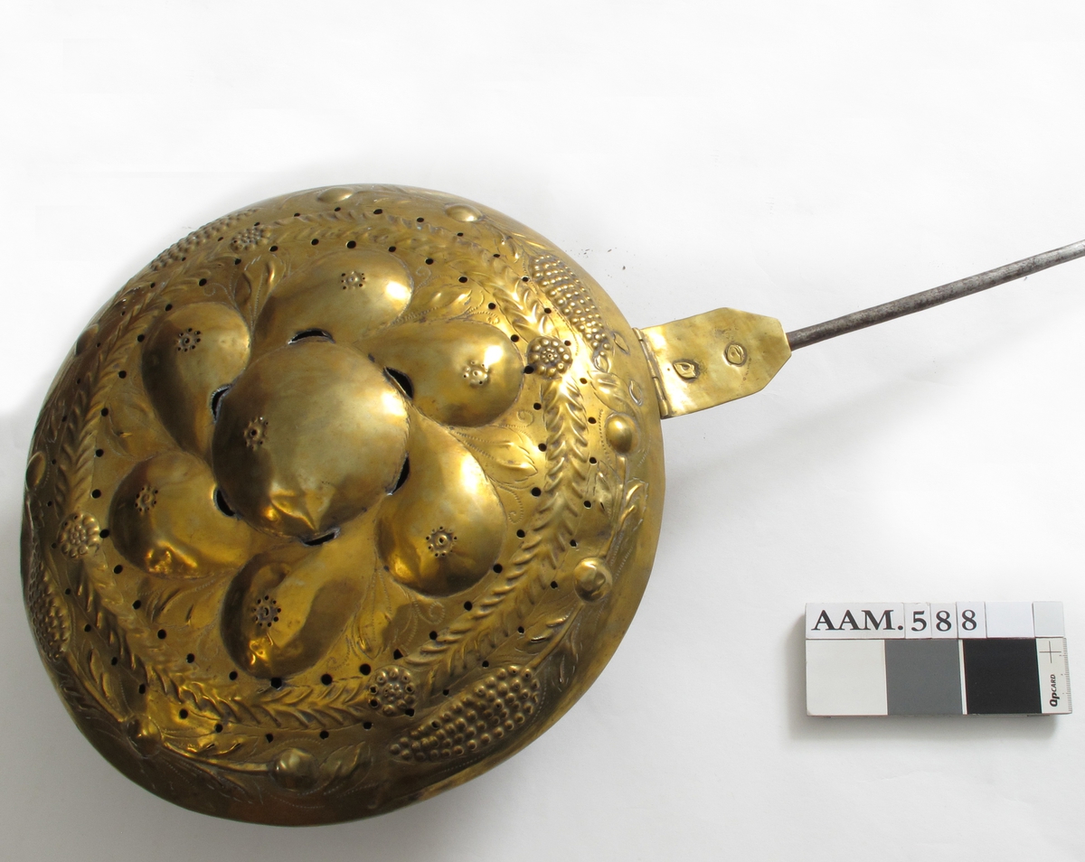 Sengevarmer.   messing.   Beholder rund. Lokk drevet med uthamrede ornament.  Under kanten jernring. Skaft jern.  Messing,   drevet, punslet og gjennombrutt arbeid,   innvendig fortinnet.  I lokket innvendig sees   kobber.  Midt på lokket en stor rund bule (eple?) med 6  pæreformete buler omkring, i mellomrommene mellom  pærene stiplete blad i lavere relieff. Langs kanten  en bladgren, ytterst med 4 kongleformer på gren med  blad. Sekskantet hengselbeslag med to messingnagler,  over det nederste utydelig stempel  Langt  jernskaft med to profilringer øverst.  På baksiden svakt synlige eierinitialer.  Skaftet går over i jernring rundt pannen., som har skrånende sider mot bunnen. RH.