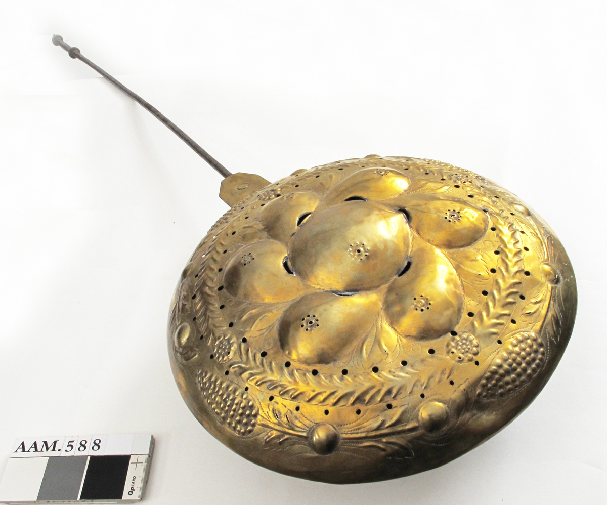 Sengevarmer.   messing.   Beholder rund. Lokk drevet med uthamrede ornament.  Under kanten jernring. Skaft jern.  Messing,   drevet, punslet og gjennombrutt arbeid,   innvendig fortinnet.  I lokket innvendig sees   kobber.  Midt på lokket en stor rund bule (eple?) med 6  pæreformete buler omkring, i mellomrommene mellom  pærene stiplete blad i lavere relieff. Langs kanten  en bladgren, ytterst med 4 kongleformer på gren med  blad. Sekskantet hengselbeslag med to messingnagler,  over det nederste utydelig stempel  Langt  jernskaft med to profilringer øverst.  På baksiden svakt synlige eierinitialer.  Skaftet går over i jernring rundt pannen., som har skrånende sider mot bunnen. RH.