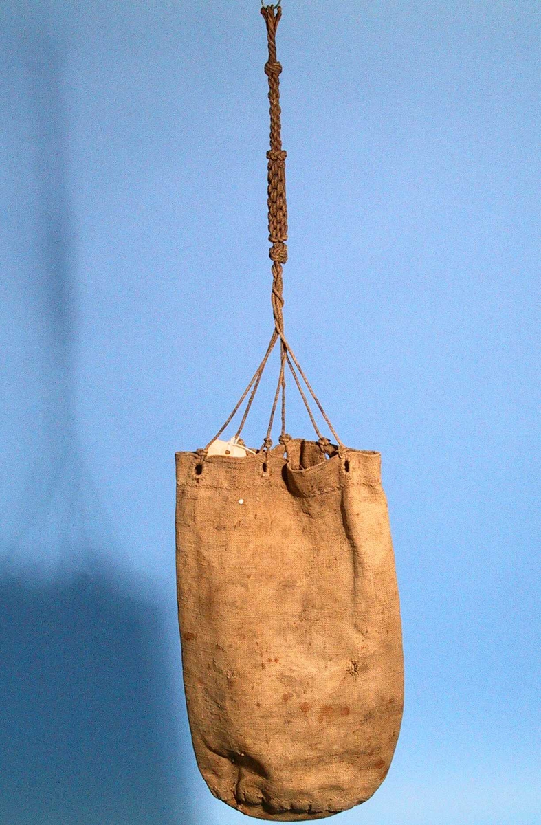 Grov brun lerret, hyssing. En søm i siden, 6 hull i kanten, sydd med store  knapphullsting. Uten  snoretrekk, men med stroppende festet i hvert hull og knyttet til  ca. 26 cm lang stropp  Tilstand mai 1967: 4 - 5 små hull i bunnen, antag. p.g.a.  merlespikeren. 