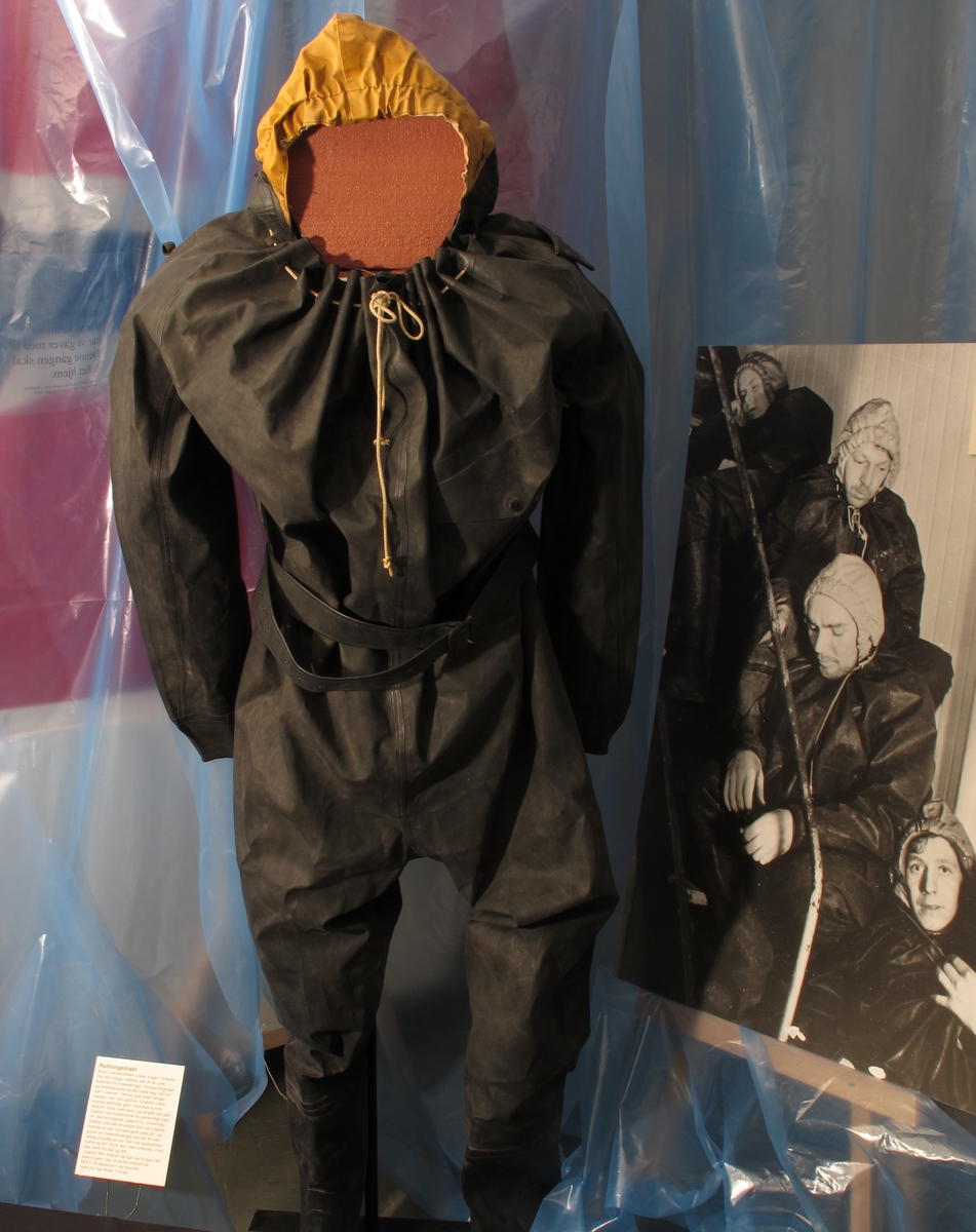 Gummidress, brukt av alliert marine og handelsflåte under krigen 1940 - 45. 
Sort dress med gul hette. Stor gummiddress med påsatte støvler, som har blyinnlegg for å holde drakten flytende loddrett. Lange ermer, vid halsåpning med snoretrekk og gul hette med innvendig hvit elastikk. Innv. bruksanvisning.  Stempel under sålen.
