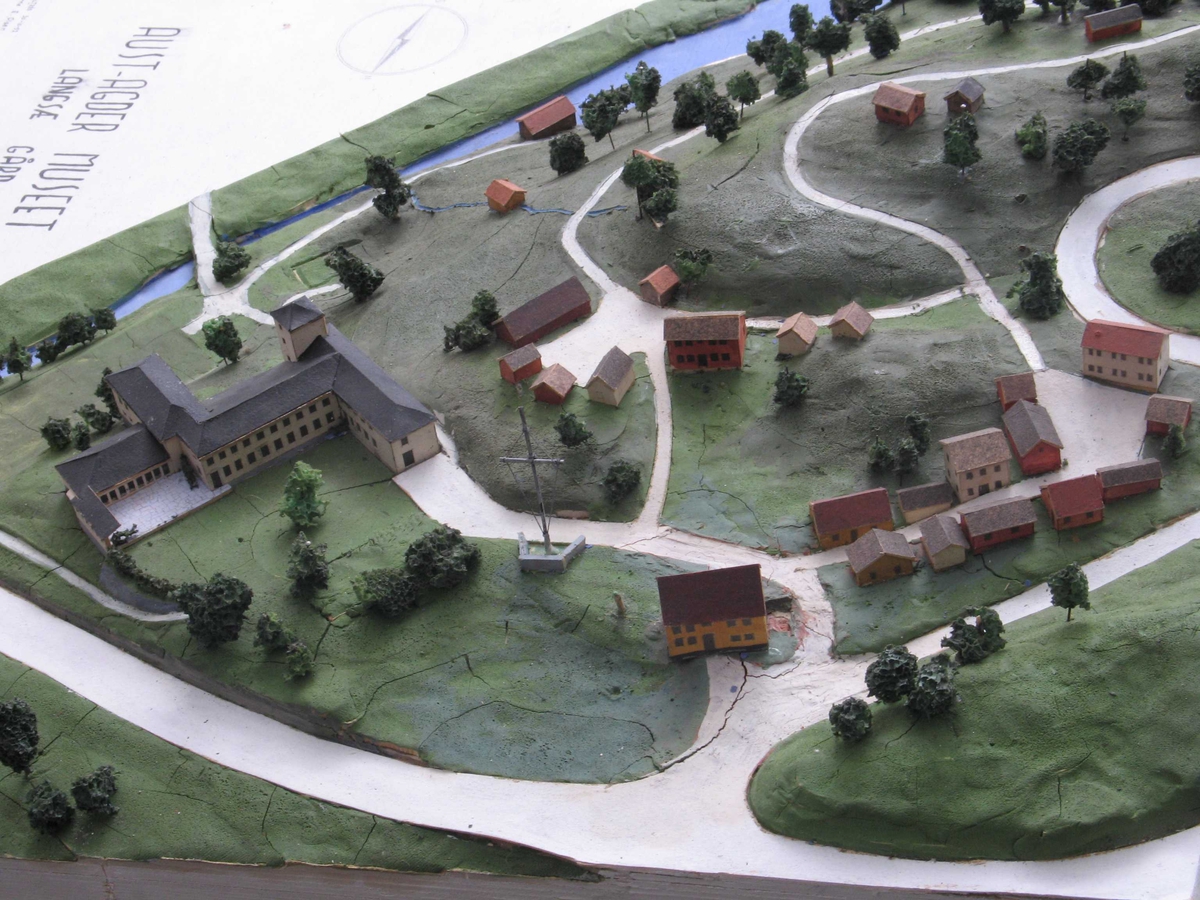 Plan og modell over Aust Agder Museet,  1952.   Furukasse  med   finerlokk,  I kassen en modell av Langsæterrenget,  med Barbuelva. Hovedbygningen fra 1858 er borte,  museumsbygget er større enn nå. Rundt i  terrenget spredte tun og bygate = Aust  Agder By og Folkemuseum (som omtalt bl. a.  i hefte 1934, Albert Uglands plan for museet).  