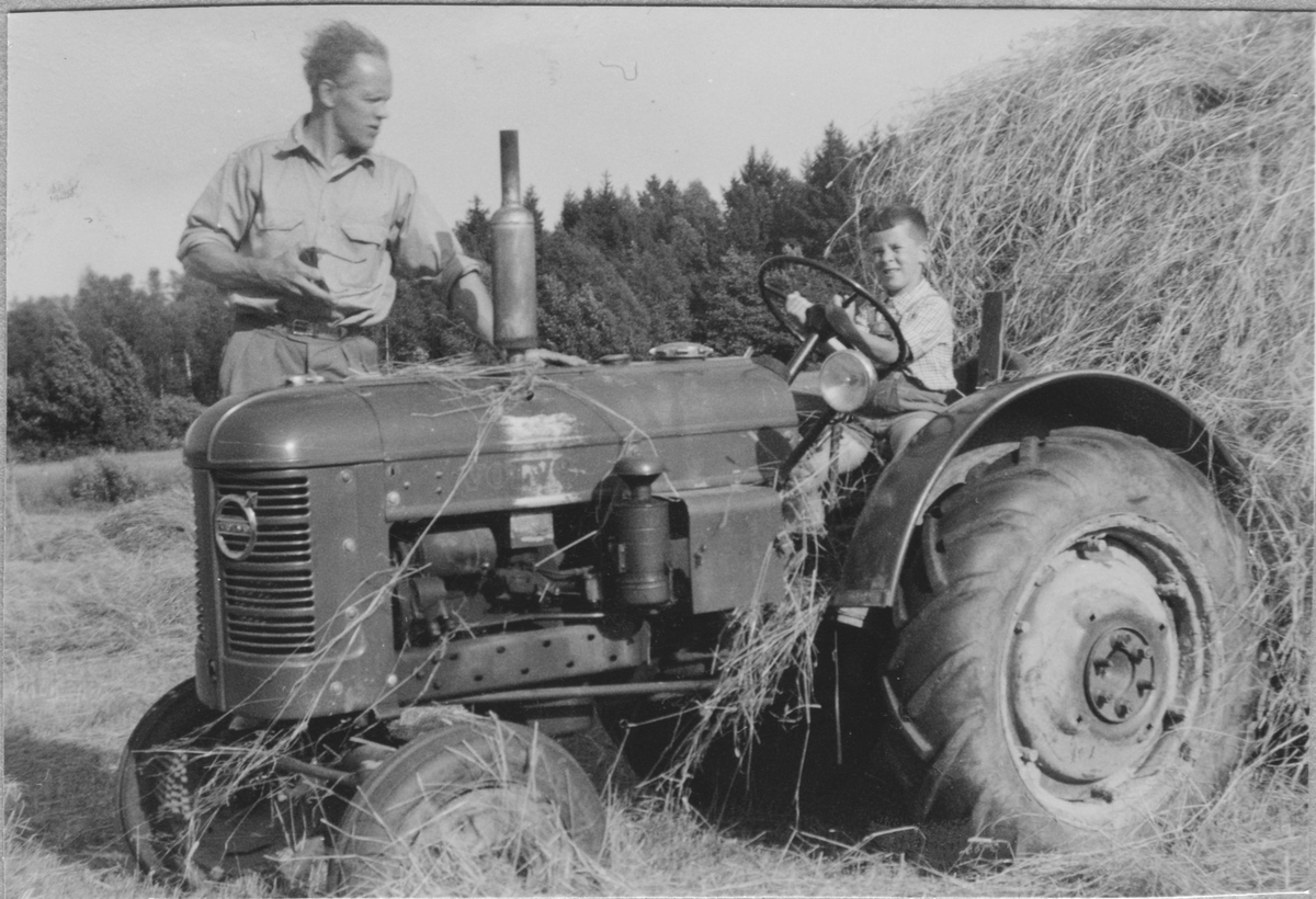 Gutt på traktor, kjører høy. Mann ved siden av.