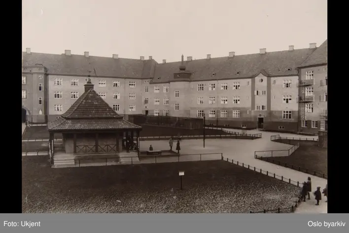 Husfasader mot indre gårdsplass. Inngjerdede plener. Barn i lek. Trafobygg. Fra kvartal VIII i Torshov-kvartalene (Torshovbyen). Sannsynligvis fra 1920-tallet. Albumtittel: "Fotografier over Kristiania kommunes boligbebyggelse".