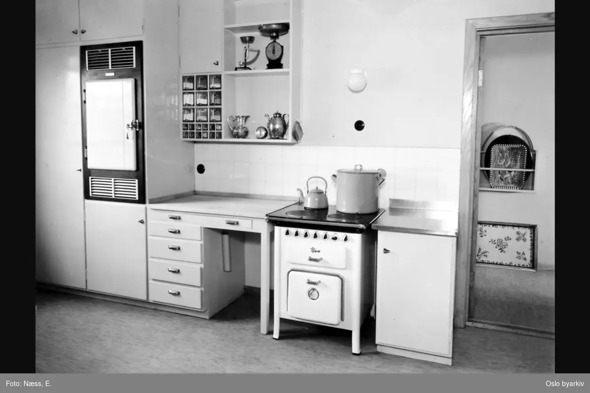Tjenestekjøkken med kjøkkenbenk, overskap, komfyr og kjøleskap. Albumtittel: "Oslo hovedbrannstasjon" (eg. identisk bildetittel til alle bildene i albumet)