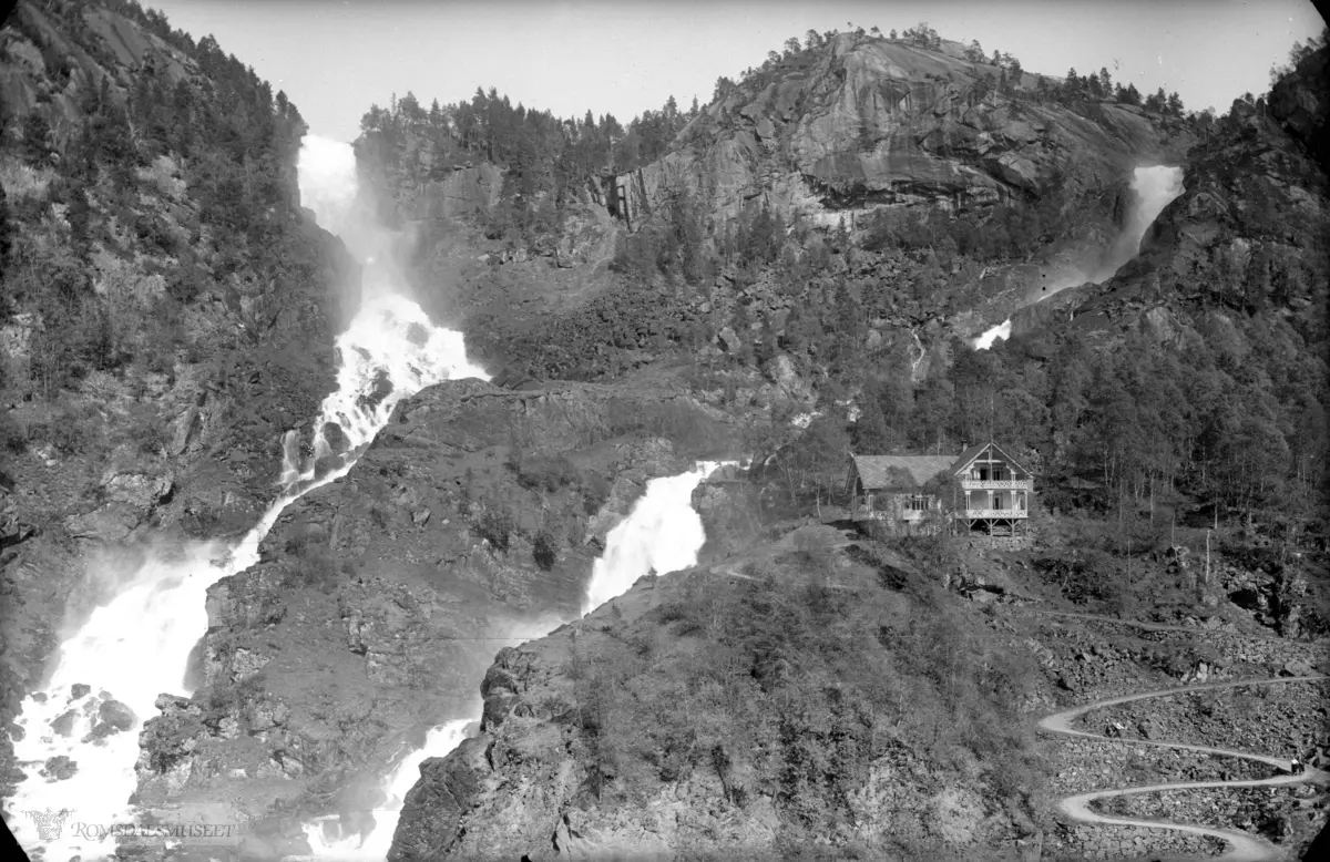 Låtefoss, hotellet og veien..Låtefoss er en 165 m høy foss i Odda, Hordaland. Den er den mest kjente av fossene i Oddadalen, som har flere fosser innenfor en strekning på 10 km..Fossen er en av Norges mest besøkte naturbaserte turistattraksjoner. Den er spesiell i det at den har to overløp fra Låtevatnet som flyter sammen midt i fossen. Den er vernet, og den har jevn vannføring året rundt..Låtefossen var et yndet reisemål for engelske og tyske turister som besøkte Odda på 1800-tallet. For å komme til fossen ble turistene skysset med hest og kjerre fra fjorden og opp langs Sandvinvatnet og videre opp Oddadalen til Låtefoss. Det var også et hotell ved fossen. I dag kan man se rester etter grunnmuren hotellet stod på, på sørsiden av fossen..Riksvei 13 krysser den nedre delen av fossen på ei steinhvelvbru.