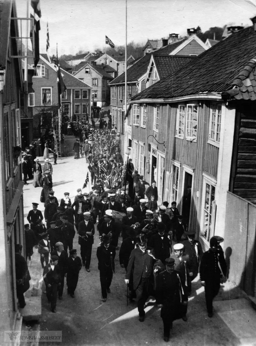 17 mai feiring i 1906, Toget passerer Torget. .Til venstre ser em litt av B ? Som ved krigsbrannen i 1940 eides av Johan Hammerø. Videre ser en litt av S.Petterssons forr.bygg. Midt på bildet i bakgrunnen storgata. M.Breckes forr.gård og litt opp i Myrabakken O.A Sannes forretnings gård. Til høyre i forgrunnen Storgata 37 (341)( (Høstmarkgården). Videre ser en i gavlen av O.Løchens forr.går og deretter bakhandler Dahls forr gård.