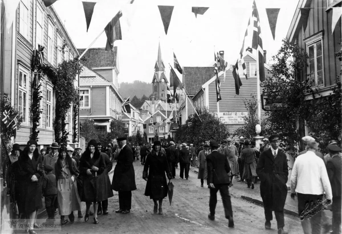 Storgata sett østover., Landbruksutstillinga 1931..Molde by ferdig til ta i mot Kongen. Kirken i bakgrunnen, Kildal & Co til høyre. "Storgata var pynta til fest og det er fullt av folk som gjerne ville ha eit glimt av kong Haakon. . .Blikkenslager Jakob Jacobsen f.1857 (Blik & Kobberslager).