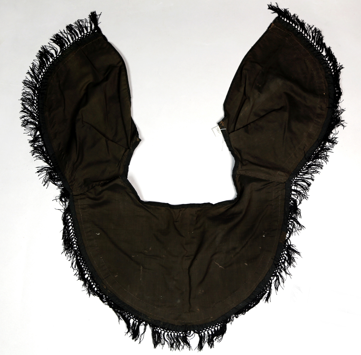 Krage som henger over skuldrene og er rund i formen. Den har fløyelsbånd og i nedkant bånd med frynser. Festes med hekter. 