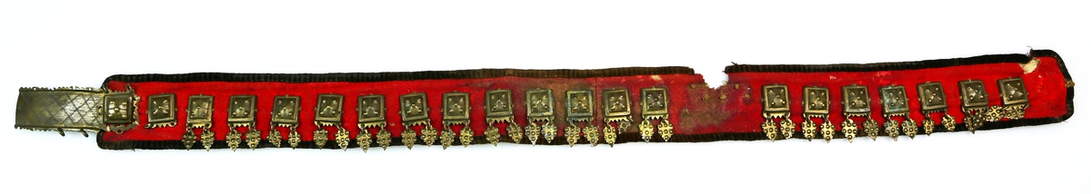 Stølebelte av rødt ullstoff kantet med mørkt ullstoff og med striestoff på innsida. Det er små beltestøler langs hele beltet, som hver har to lauv. Beltespenna har enkel gravert dekor. 