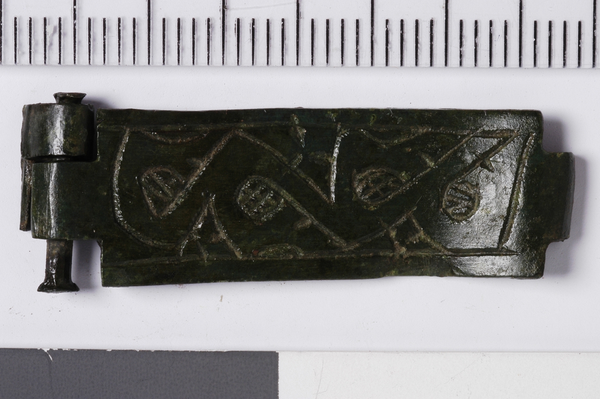MÅL: L. 3, 8 CM B. 1, 2 CM, REST AV HENGSEL I ENE ENDEN. REKTANGULÆR PLATE SOM ENDER I EN OMBØYD HEKTE. PLATEN HAR FLETTVERKS ORNAMENTIKK.