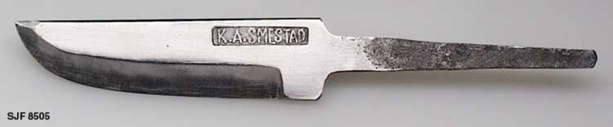 Knivblad, smidd av smeden Knut A. Smestad (1906-1995) fra Åmot i Modum i Buskerud. Gjenstanden er 14,7 centimeter lang. Sjølve bladet er 7,8 centimeter langt og det er 2,1 centimeter bredt ved brystningen. Det er cirka 4 millimeter tjukt ved brystningen og tynnes noe mot spissen. Bladryggen er rett. Også den bakre delen av eggen er rett mens den fremre er konvekst buet mot bladspissen. Bladet har cirka 9 millimeter brede skipefaser på begge sider. Smedens navnestempel er plassert øverst på den bakre delen av venstre bladside. Bladet og den fremre delen av tangen er blankpolert. Den fremre enden av tangen er om lag 1,0 centimeter bred, den bakre enden bare halvparten så bred. 

Kniven er smidd i 16 millimeters firkantjern hvor det er slått ut en spalte i jernet. Knut lærte smiing av faren Asle Knutsen Smestad (1871-1958). «AKS» på bladene Knut laget stempelet til faren og seg selv i 1924.