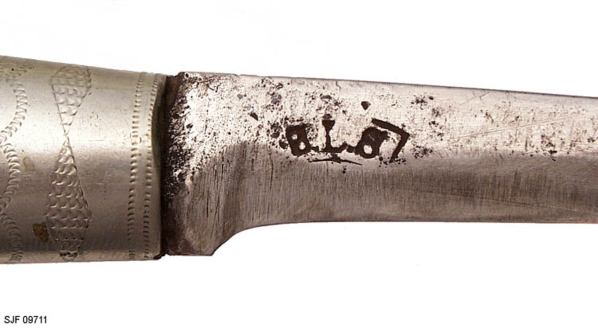 Tollekniv med slire.  Slira er signer "T. B. ", som skal være initialene til Trygve Brenna fra Åmot på Modum.  Bladet er 8, 2 centimeter langt og 1, 9 centimeter bredt ved brystinga.  På den bakre delen av nakken, som er om lag 0, 5 centimeter bred, er det filt to tverrstreker og et kryss.  Høyre bladside har tre innstemplete initialer, antakelig "Ø. L. S. ", innskrevet i ei buet ramme.  Denne signaturen ble brukt av eggsmeden Knut Knutsen Øyo (1860-1921), og seinere av firmaet til sønnene Lars, Thomas, Knut og Olav Øyo, «Øyo ljåsmie».  Dette navnet ble brukt etter 1. verdenskrig.  Firmanavnet ble for øvrig endret til «Brødrene Øyo» i 1941, som en konsekvens av at produktspekteret bedriften kunne levere var blitt bredere, og av at ljåblader ikke lenger var det dominerende produktet.  Bladet på denne kniven bærer preg av å være slipt mange ganger. 

Skaftet er lagd av flammebjørk Det er snaut 9, 5 centimeter langt og har en noe konveks form i lengderetningen og et ovalt tverrsnitt (snaut 2, 5 X 1, 95 centimeter på midten).  Begge ytterender er forsterket med 2, 0 centimeter brede nysølvholker med inngraverte linje-, bue- og rombemønstre.  I enden av bakholken er det påsatt ei rombeformet plate, som den dreide knoppen enden av knivbladets tange er forankret i er festet mot.  Tredelen av skaftet har en lys gulbrun farge som muligens skyldes forsiktig beising.  Kniven - inkludert blad, skaft og knopp - er 18, 7 centimeter lang.  

Kniven har treslire, lagd av flammebjørk og bundet sammen av nysølvbeslag.  Slirebeslaget består av to spenger langs sidene, som holdes ihop av tre holker: en munningsholk (øverholken), en midtholk og en doppsko.  Munningsholken har samme dekor som holkene på skaftet, altså linjemønster (parallelt med overkant), bue- og rombemønster (skravert).  Nederkanten på denne holken har et profilert mønster på den sida som skulle vendes fram når kniven ble båret i bukselinninga.  På baksida er det påloddetei hengsle for en beltestropp med "smikk" av brunsvart lær.  Den bakre delen av denne stroppen har spalte for på- og avknepping, den fremre et hull for forankring av kniven i knoppen i skaftenden.  Midtholken på slira har inngravert to "skraverte" romber, og har profilerte kanter på forsida av slira.  Baksida er glatt, bortsett fra at det er her knivmakeren har gravert inn initialene sine med ei kort pil på hver side.  Doppskolen er tilnærmet rektangulær og har en trefliket "rombe-rosett" som en profilert overkant som dekor på forsida.  I enden av doppskoen er det påsatt en dreid knopp av samme type som den som er brukt i øverenden av skaftet. 

Redskapet veier 135 gram, herav sjølve kniven 70 gram. 