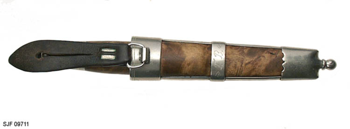 Tollekniv med slire.  Slira er signer "T. B. ", som skal være initialene til Trygve Brenna fra Åmot på Modum.  Bladet er 8, 2 centimeter langt og 1, 9 centimeter bredt ved brystinga.  På den bakre delen av nakken, som er om lag 0, 5 centimeter bred, er det filt to tverrstreker og et kryss.  Høyre bladside har tre innstemplete initialer, antakelig "Ø. L. S. ", innskrevet i ei buet ramme.  Denne signaturen ble brukt av eggsmeden Knut Knutsen Øyo (1860-1921), og seinere av firmaet til sønnene Lars, Thomas, Knut og Olav Øyo, «Øyo ljåsmie».  Dette navnet ble brukt etter 1. verdenskrig.  Firmanavnet ble for øvrig endret til «Brødrene Øyo» i 1941, som en konsekvens av at produktspekteret bedriften kunne levere var blitt bredere, og av at ljåblader ikke lenger var det dominerende produktet.  Bladet på denne kniven bærer preg av å være slipt mange ganger. 

Skaftet er lagd av flammebjørk Det er snaut 9, 5 centimeter langt og har en noe konveks form i lengderetningen og et ovalt tverrsnitt (snaut 2, 5 X 1, 95 centimeter på midten).  Begge ytterender er forsterket med 2, 0 centimeter brede nysølvholker med inngraverte linje-, bue- og rombemønstre.  I enden av bakholken er det påsatt ei rombeformet plate, som den dreide knoppen enden av knivbladets tange er forankret i er festet mot.  Tredelen av skaftet har en lys gulbrun farge som muligens skyldes forsiktig beising.  Kniven - inkludert blad, skaft og knopp - er 18, 7 centimeter lang.  

Kniven har treslire, lagd av flammebjørk og bundet sammen av nysølvbeslag.  Slirebeslaget består av to spenger langs sidene, som holdes ihop av tre holker: en munningsholk (øverholken), en midtholk og en doppsko.  Munningsholken har samme dekor som holkene på skaftet, altså linjemønster (parallelt med overkant), bue- og rombemønster (skravert).  Nederkanten på denne holken har et profilert mønster på den sida som skulle vendes fram når kniven ble båret i bukselinninga.  På baksida er det påloddetei hengsle for en beltestropp med "smikk" av brunsvart lær.  Den bakre delen av denne stroppen har spalte for på- og avknepping, den fremre et hull for forankring av kniven i knoppen i skaftenden.  Midtholken på slira har inngravert to "skraverte" romber, og har profilerte kanter på forsida av slira.  Baksida er glatt, bortsett fra at det er her knivmakeren har gravert inn initialene sine med ei kort pil på hver side.  Doppskolen er tilnærmet rektangulær og har en trefliket "rombe-rosett" som en profilert overkant som dekor på forsida.  I enden av doppskoen er det påsatt en dreid knopp av samme type som den som er brukt i øverenden av skaftet. 

Redskapet veier 135 gram, herav sjølve kniven 70 gram. 