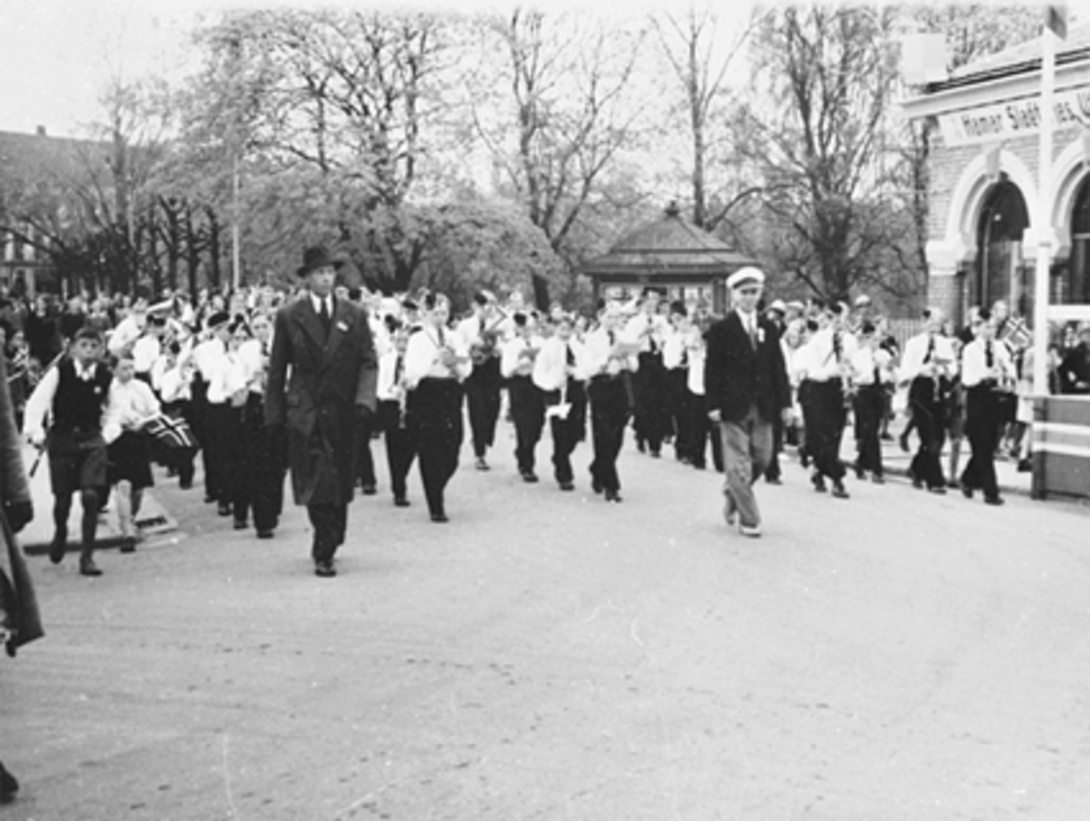 17 MAI TOGET FORAN BASARBYGNINGEN MED HAMAR GUTTEMUSIKKORPS MED DIRIGENT HALFDAN HANSEN, KIOSKEN I STRANDGATEPARKEN I BAKGRUNNEN