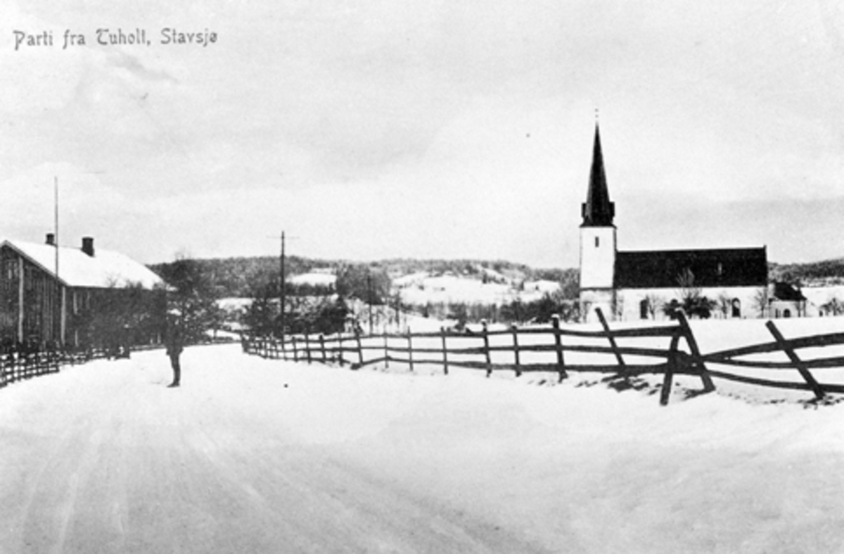 Postkort. "Parti ved Tuholt, Stavsjø". Til venstre ligger Jevne landhandel. Stavsjø kirke. Vinter. Gjerde langs vegen. Navnet Tuholt er ukjent og trolig feil.