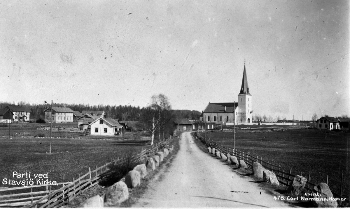 Postkort "Partie ved Stavsjø Kirke". Stabbesteiner og gjerde langs vegen. Til venstre er den eldste og den nye skolebygningen ved Penningrud. Småbruket Arneberg. Stallene ved kirken og Kirkenær landhandel. Trolig før 1924.