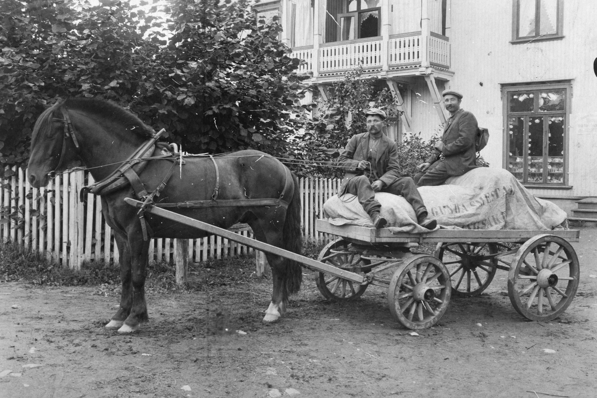 Hest og kjerre, varetransport. Presenning merket "Gunda Smedstad, Ringsaker. " Til  venstre Lars "Eriksrud" Mathiasen og ukjent kjører varer for forretningen i Eriksrud innehaver Smedstad. Bildet er tatt utenfor forretningen til Ludv. Larsen som hadde forretning i huset ved Nedre Bruvold (Sundgaard). Ca. 1920-1930.