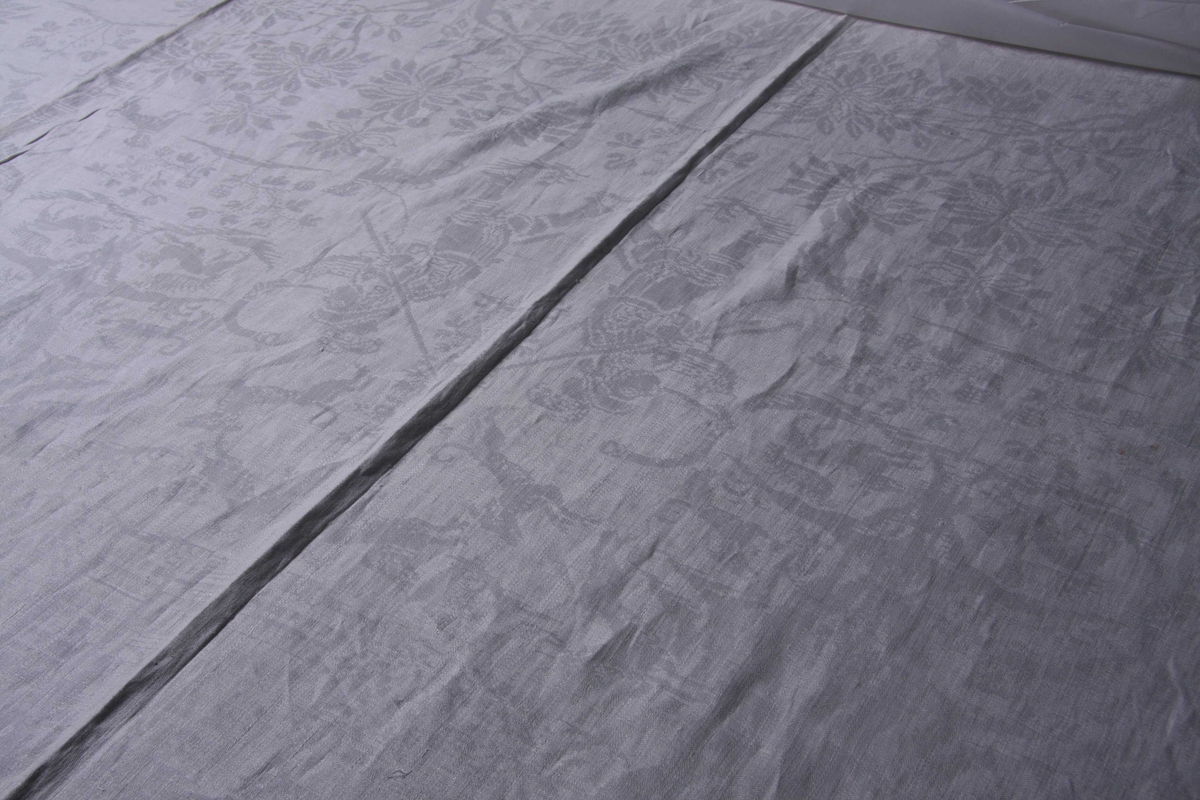 Hvit lindamask med sjakkbrettbord langs sidene og bord innenfor av motstillte blomsterranker og fugler. På dukens midtparti består mønsteret av parvise motstillte figurer i følgende rekkefølge fra den ene enden. Damer til hest med jaktfalk. Jegere med hellebard og jakthorn omgitt av hunder som forfølger hjort i tregruppe.Mellom trekronene sees jaktslott. Jeger med gevær i anlegg. Fontene med svømmende og flyvende fugler og liggende enhjørninger. innimellom enkelte av figurradene er det store blomstermotiver. Mønsteret gjentaes speilvendt på høyre og venstre side av duken.