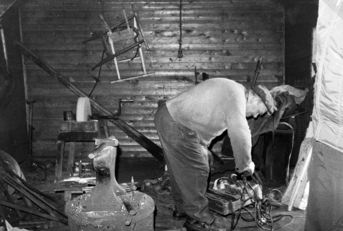 Interiør, smie. Erik Granberg f. 1912 i smia si på Haugli på Tangen. På veggen bak ses et par greip, treverket av to sparkstøttinger, pluss flattjern som skal brukes til meier på disse. En slipestein, ambolt og hammere pluss smeden sjøl i kjent positur. 