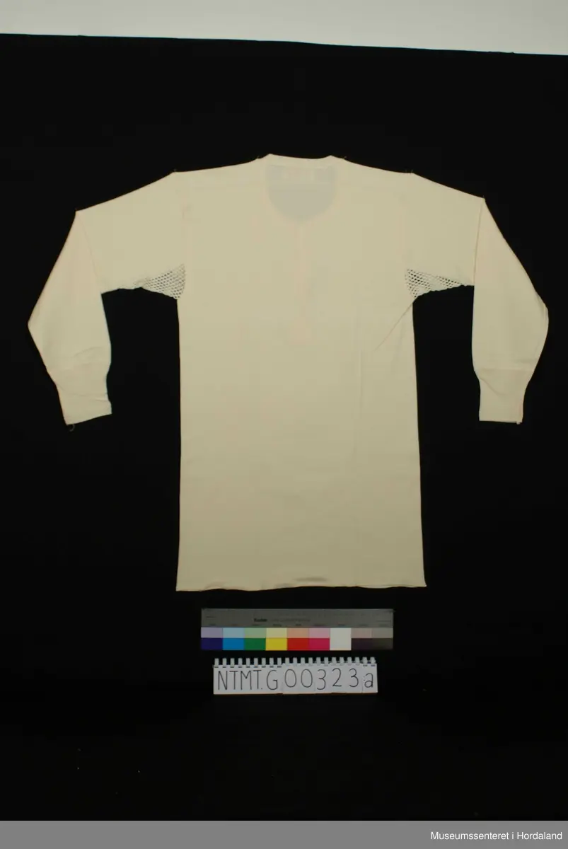 Rund hals med åpning midt foran med to knapper, lange ermer med brotavslutning, kile under ermer i helsetrøyestoff, skulderstykker.