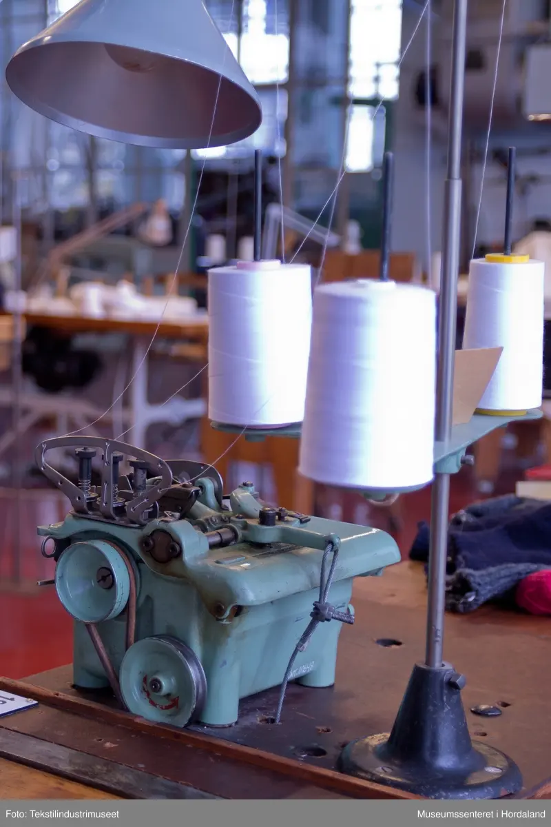 Mingrøn 1-nåls overlockmaskin som syr saman to delar trikotstoff, og samtidig kuttar av overflødig stoff med ein kniv. Maskina har tre trådar, men ein kan òg tre den med to trådar. Den har ei pumpe som oljar maskina automatisk.

Maskina er montert på eit Singer-bord saman med fleire andre symaskiner etter at den kom til museet. Bordet har eit hol der kappet frå symaskinene skulle dette ned.