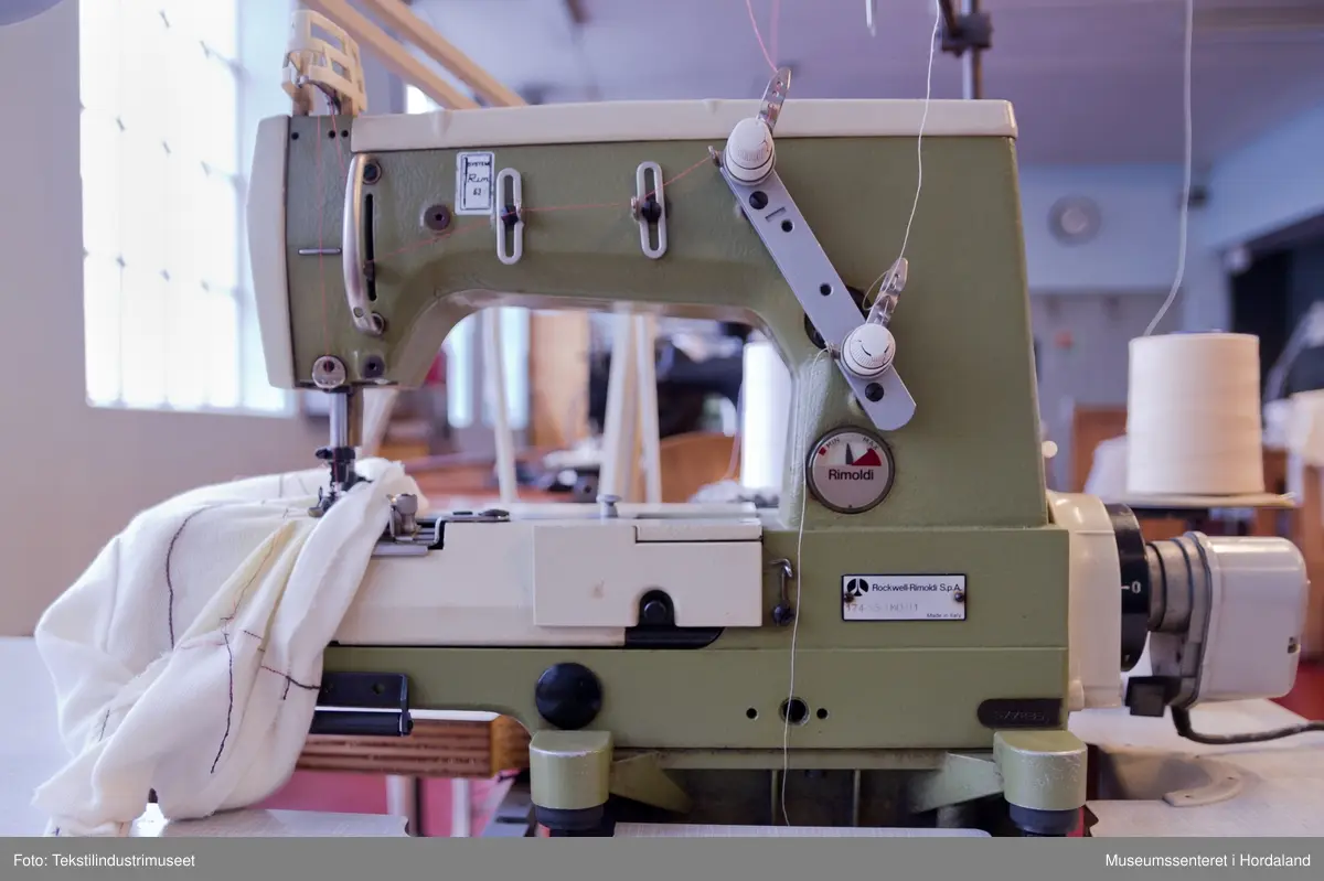 Rimoldi-symaskin montert på lite bord med pedalar og motor under bordet, og stativ til to trådsneller og trådførarar ved sidan av maskina. Strømbrytar er plassert under bordet.

Motor: Etka Variostop
Typ: VD 12
Nr. 08315789
3~Mot (triangel)Y 220/380 V
2,6 / 1,5 A     550 W
2800 / 3400 1/min
...
Made in Western Germany