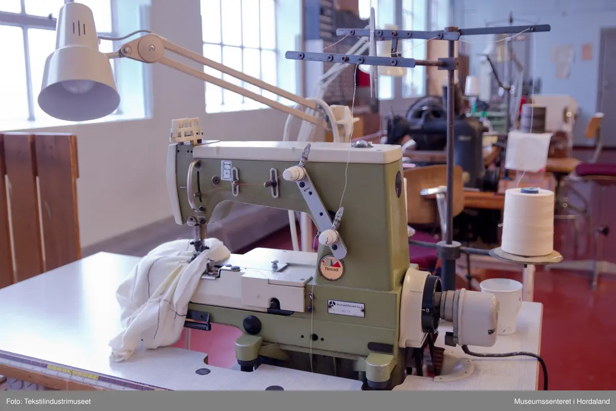 Rimoldi-symaskin montert på lite bord med pedalar og motor under bordet, og stativ til to trådsneller og trådførarar ved sidan av maskina. Strømbrytar er plassert under bordet.

Motor: Etka Variostop
Typ: VD 12
Nr. 08315789
3~Mot (triangel)Y 220/380 V
2,6 / 1,5 A     550 W
2800 / 3400 1/min
...
Made in Western Germany