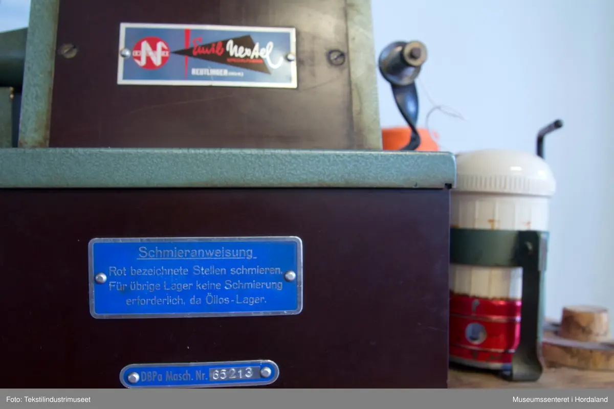 Grøn og brun maskin som lagar duskar/pompongar. Maskina har trådstativ med seks trådførarar, med ein kvit bomullstråd inni dusken og teljeapparat montert på toppen. Den står på eit Singer-bord, og av-på knappen er fastmontert i sjølve bordet.