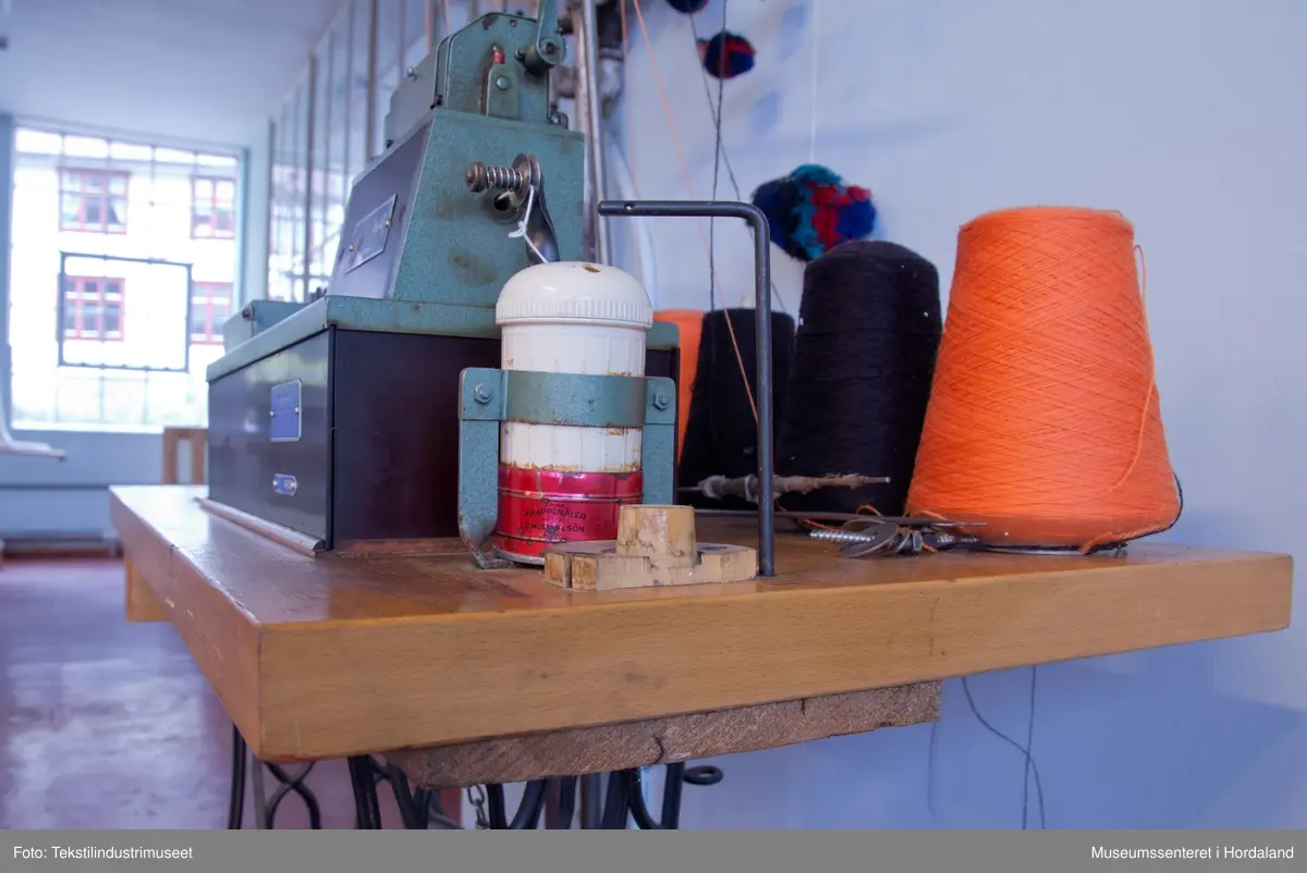 Grøn og brun maskin som lagar duskar/pompongar. Maskina har trådstativ med seks trådførarar, med ein kvit bomullstråd inni dusken og teljeapparat montert på toppen. Den står på eit Singer-bord, og av-på knappen er fastmontert i sjølve bordet.