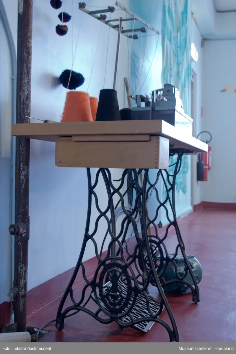 Grøn og brun maskin som lagar duskar/pompongar. Maskina har trådstativ med seks trådførarar, med ein kvit bomullstråd inni dusken og teljeapparat montert på toppen. Den står på eit Singer-bord, og av-på knappen er fastmontert i sjølve bordet.