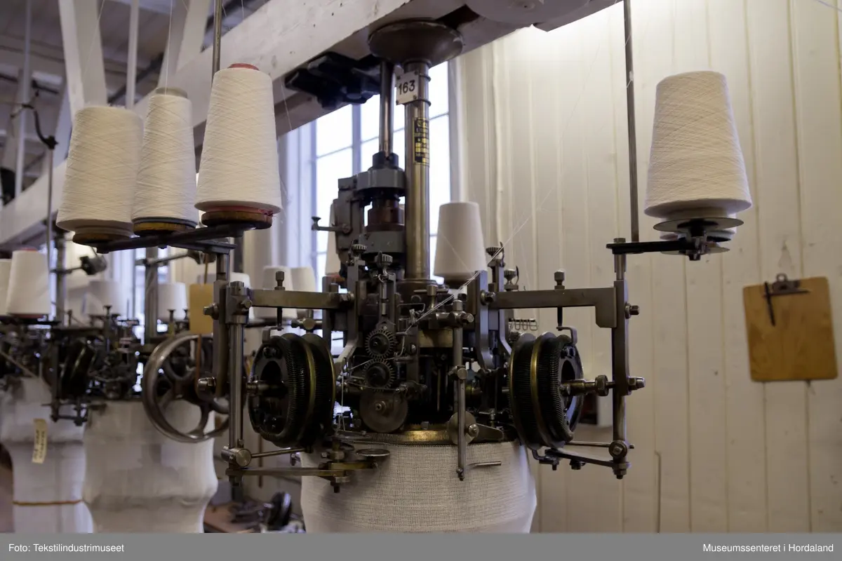 Rundstrikkemaskin/rundstol som er montert med eit feste og er kopla til reimdrift i taket. Maskina har tre system og ei nedtrekksmekanisme nederst med små metallpiggar montert på tekstilt materiale.