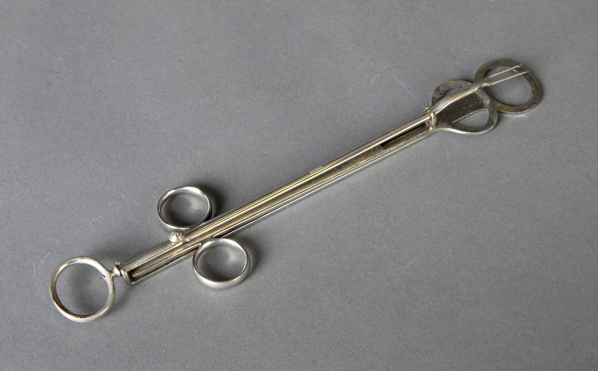 Metallstang som ender i 2 spisser med mothake, to flate metallstenger som ligger mot hverandre og ender i hver sin ovale kniv. 3 ringer til fingertak.
