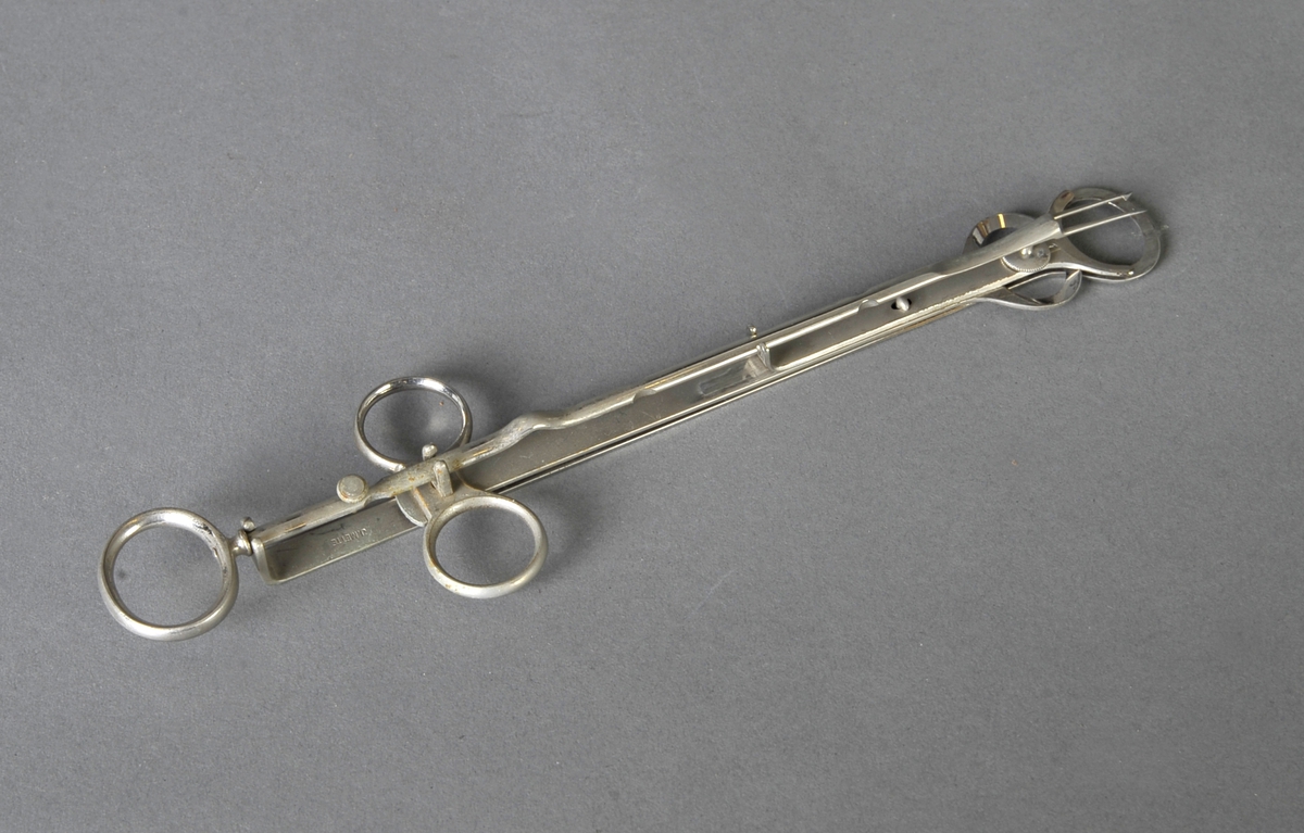 Metallstang som ender i 2 spisser med mothake, to flate metallstenger som ligger mot hverandre, er bevegelige og ender i hver sin ovale kniv. 3 ringer til fingertak.
