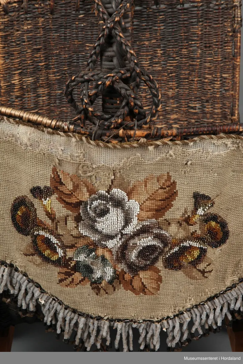 Fletta korg med utskårne trebein. Korga har ein u-form med to opne sider. Handtaket er forsterka med ei trespile. Handtaket har fletta band som ekstra dekorasjon langs heile handtaket. På eine sida (framsida) er det festa eit broderi til korga som er brodert med halve korssting og perler - rosemotiv. Broderiet er fora med eit mørkegrønt? stoff og har i tillegg eit tøystykke inni. Broderiet har ei tvinna snor rundt kanten, bortsett frå nedre kant der det er perletredde (røyrperler) løkkjer som ein frynse-effekt.