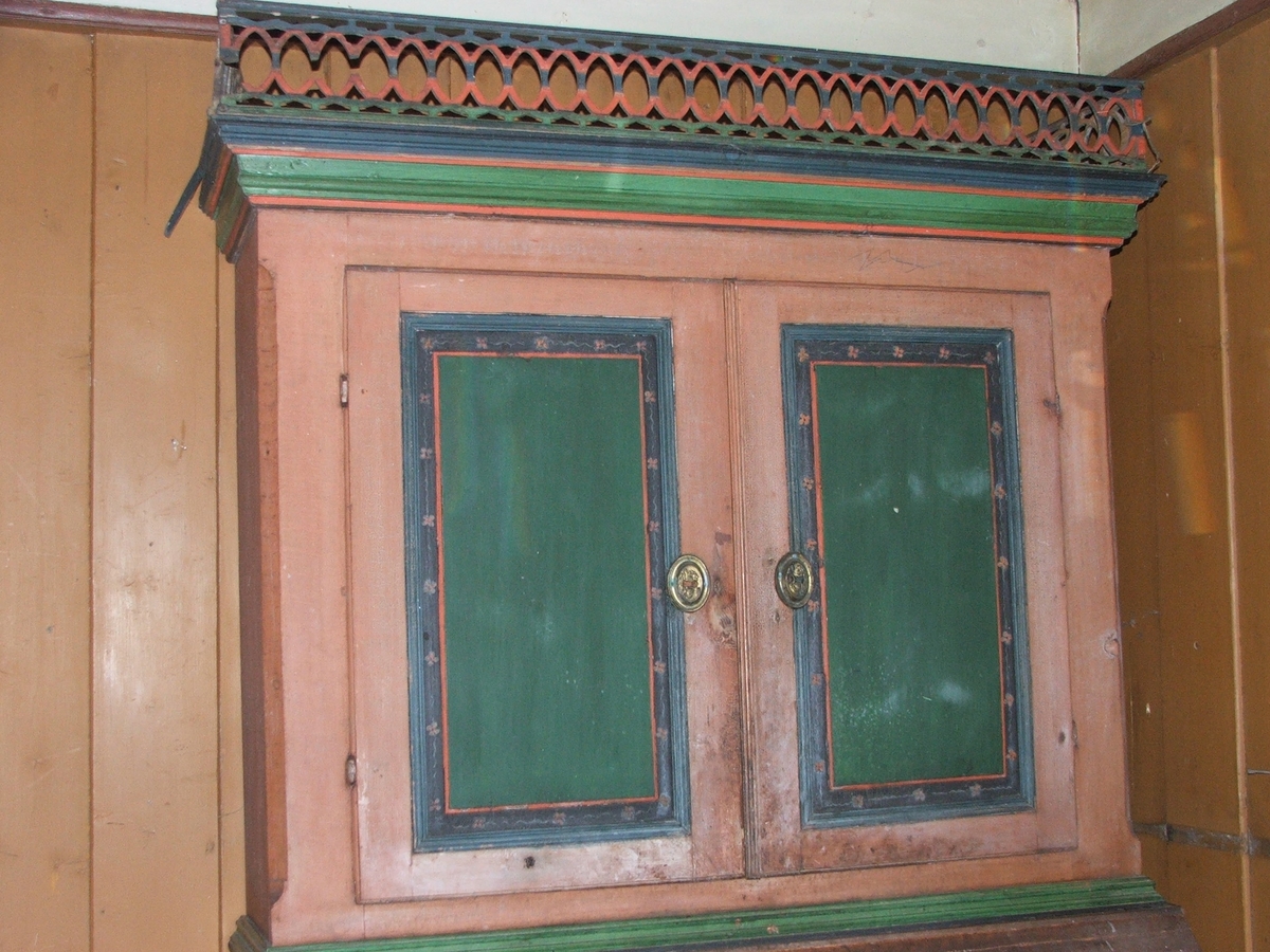 Todelt skap i tre. To skapdører øverst, 8 små skuffer i midten og fire større skuffer nederst. Oppheng til skjeer i øverste skap. Utskjæringer i tre øverst. Grønnmalte fyllinger dekorert med små blomster.