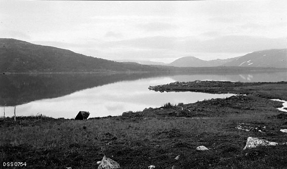 Lille Umeåvatn ved grensa mellom Mo i Rana og Sverige.  Fotografiet er tatt fra ei grasbevokst strand hvor det ligger en robåt mot et blankt vannspeil.  I bakgrunnen til venstre en skogkledd ås, til høyre en fjellrygg med snøflekker. 