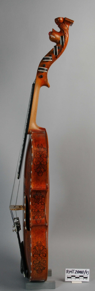 Instrument med fire strenger, og fire understrenger. Rikt dekorert. Halsen ender i et utskåret hode.