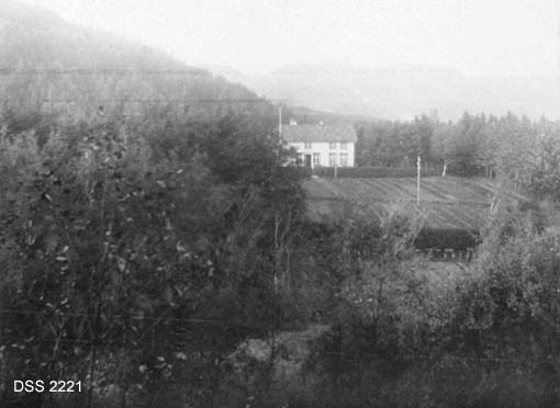 Kyrrefjord planteskole ved Farsund.  Blandingsskog i forgrunnen, kvit bygning med åkrer med plantesenger sentralt i bildeflata. 