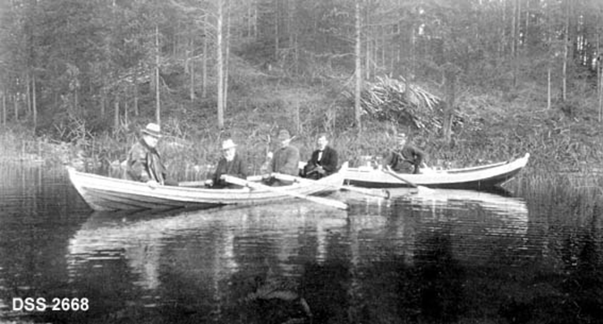 To robåter med til sammen fem mann ved skogkledd strand. Fra Einavatnet på Vestre Toten, antakelig befaring i Prestmarka. Skogen i bakgrunnen domineres av gran. En del rajeved ligger på land.