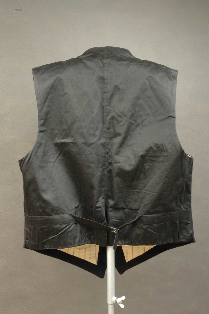 Dress i svart klede, bestående av sid jakke, vest og bukser.
Vesten har naturfarget fôr med blå striper. Fire lommer foran. Enkeltspent med fem svarte knapper. Rygg av svart, glatt bomullsstoff med spensel med metallspenne. 
Jakken har splitt bak og jakkeslag. Enkeltspent med to svarte knapper. En brystlomme på venstre side. Lukking med to knapper. Kypervevd bomullfór i bolen og blåstripete bomullsfór i ermene.
Buksa har gylf som lukkes med fire knapper og hekte. Lommer i hver side og baklomme på høyre side. Spensel midt bak. Øverst på linning er to knapper midt bak og to på hver side framme.