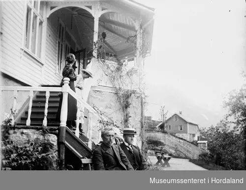 Velkledde karar på benk, kvinnar i trappa utenfor ein villa