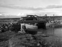 En mann sitter ved ei lita elv, en bil står på ei bru. Nysko