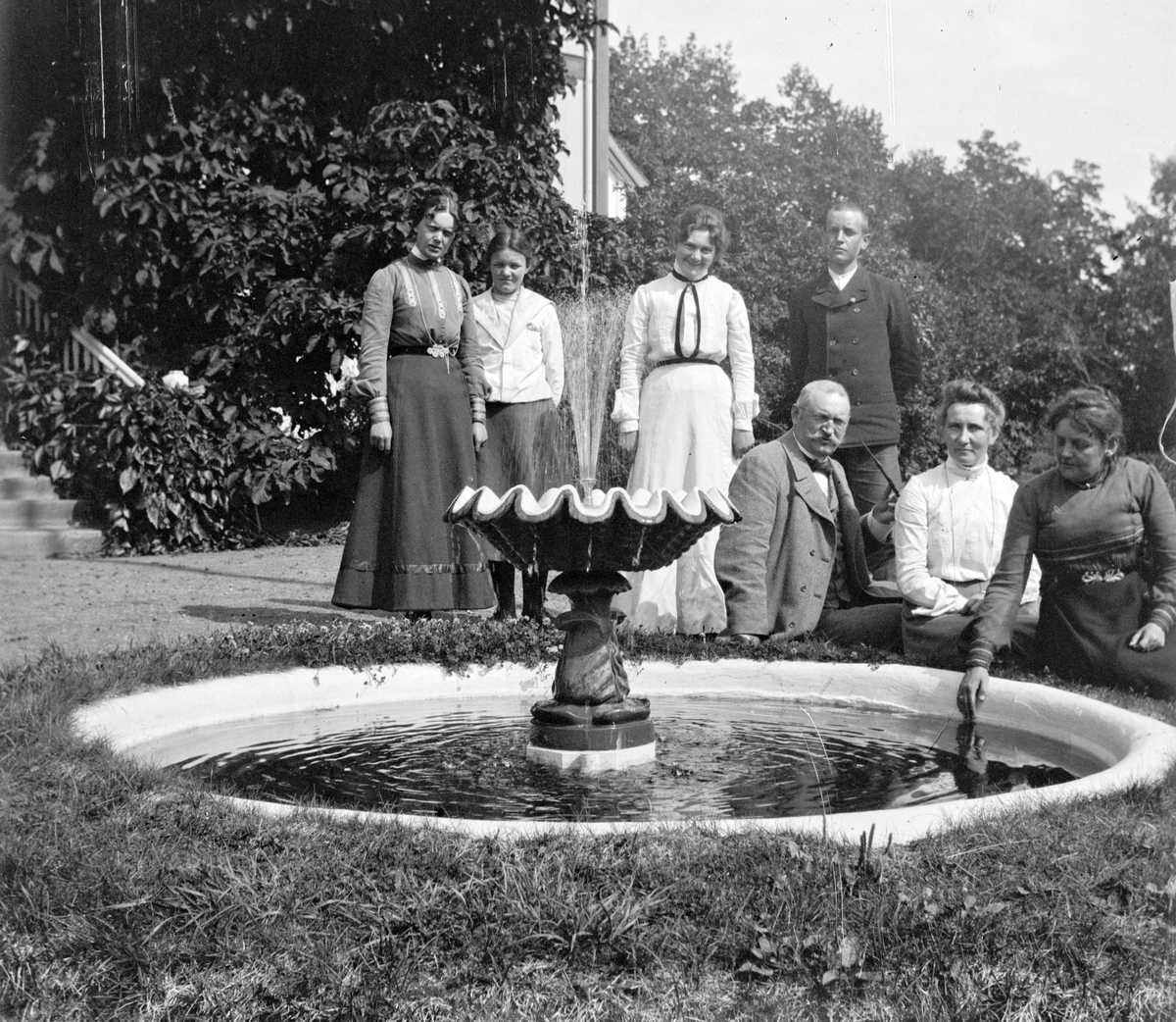 Familiegruppe ved fontenen, springvannet, ved Hovelsrud gård, Helgøya i Ringsaker. Foto Jacob Hoel.
Fra v. bak: Leonore Olssøn, Marianne Olssøn, Ragnhild Hoel, Karl Arvid Olssøn.
Sittende fra v.: Dr.Paul August Hoel, ukjent, Louise Hoel, senere gift Olssøn.
