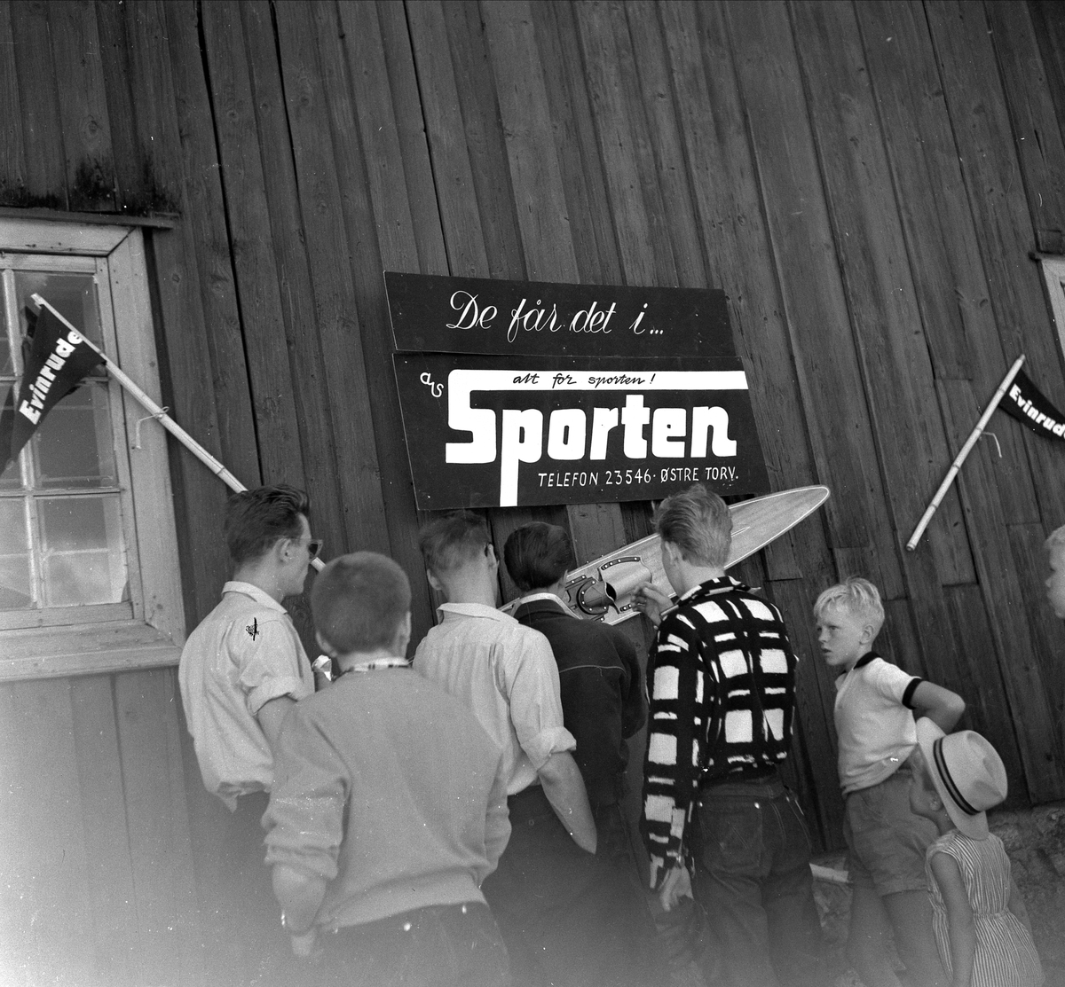 A/S SPORTENS STAND PÅ DOMKIRKEODDEN, PUBLIKUM SER PÅ BÅTMOTOR. SKILT PÅ LØTENBYGNINGEN " DE FÅR DET I AS SPORTEN" GUTTER SER PÅ VANNSKI