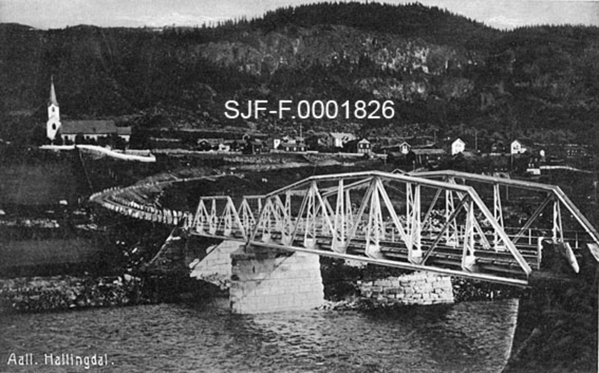 Kommunesenteret Sundre i Ål i Hallingdal (i Buskerud).  Fotografiet er tatt fra vestsida av Hallingdalsåne, fra en posisjon like nordvest for Ål jernbanestasjon på Vaterland.  Forgrunnen domineres av ei jernbru på kar av hoggen naturstein.  Denne brua førte over til jernbanestasjonen, som ble åpnet i 1907.  Stasjonsbrua var antakelig forholdsvis ny da dette fotografiet ble tatt.  Fra brua fører en veg med stabbesteiner langs vegskuldrene opp mot det daværende kommunesenteret.  Her ser vi kirka fra 1880 samt en del bebyggelse, blant annet Tingstugu (skal være fra 1767), Gretestugu (fra samme tid, ble flyttet til Kristiania) og bygninger som huset forretninger og overnattingssteder. Neden for bebyggelsen ligger sundrejordet.  Bakenfor ser vi en bratt bergskrent med barskog på toppen (Hovdene). 

Kommunesenteret Sundre ble rammet av en brann 11. mars 1931.  Brannen ødela Tingstugu, Sundre hotell og flere av forretningene som skimtes på dette fotografiet.  Stasjonsbrua i forgrunnen eksisterte fram til midten av 1960-åra, da den måtte vike plassen i forbindelse med bygginga av en ny trasé for riksveg 7 i området nede ved elva (1964-66). 