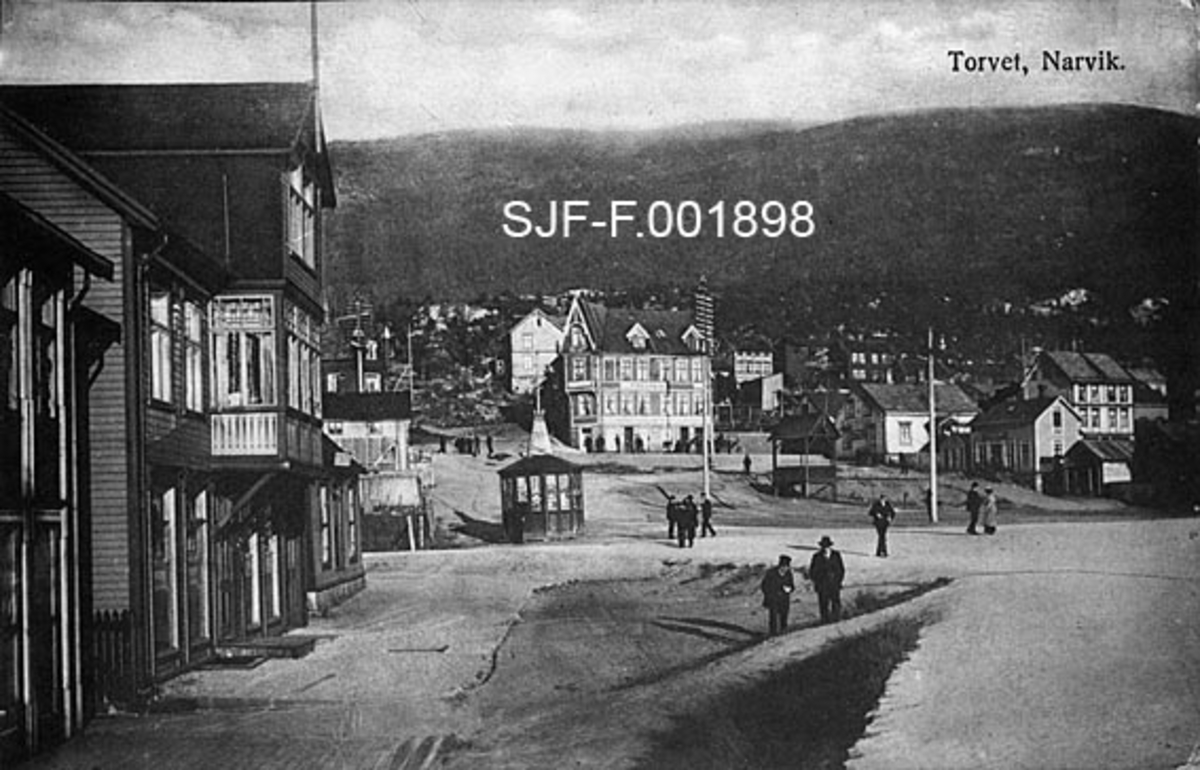 Bybilde fra Narvik i Nordland, antakelig omkring 1920.  Fotografiet er tatt over torget, som ser ut til å ha vært en stor, men nokså ujamn plass, omgitt av to-tre etasjes bolig- og forretningsgårder i tre.  En del personer, overveiende menn, spaserer på torget.  I et hjørne står en liten mangekantet bygning med ei lita flaggstang fra et tårn midt på taket.  Dette er muligens en kiosk.  I en skråning litt lengre til høyre står en stolpebåren konstruksjon med saltak, kanskje brukt som musikkpaviljong på festdager.  I bakgrunnen ei fjellside.  