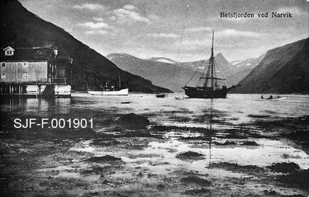 Landskapsbilde fra Beisfjorden ved Narvik i Nordland.  Beisfjorden ligger på sørsida av Ofotfjorden, vest for Narvik by.  Fotografen har stått i fjæra.  I forgrunnen skimter vi oppstikkende steiner og tang.  Til høyre i bildet et par brygger med sjøhus.  På vannet ligger det et par robåter og et par større båter, hvorav den største later til å være ei jakt.  Det kvite fartøyet, som ligger noe nærmere sjøhusene, ser også ut til å ha en slags rigg, men kan også ha hatt motordrift.  I bakgrunnen ser vi bratte fjell, dels med snøflekker litt oppe i høyden. 